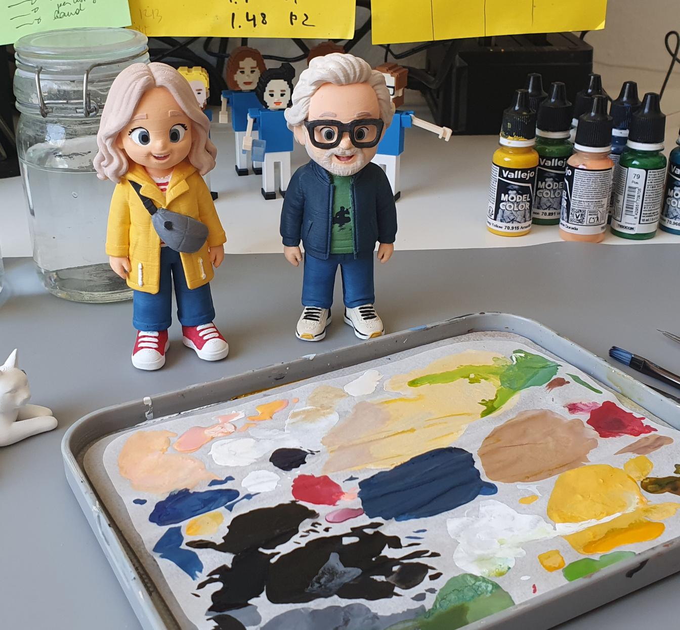 Two hand-painted RepliToy figurines on a workbench with paints and a mixing palette